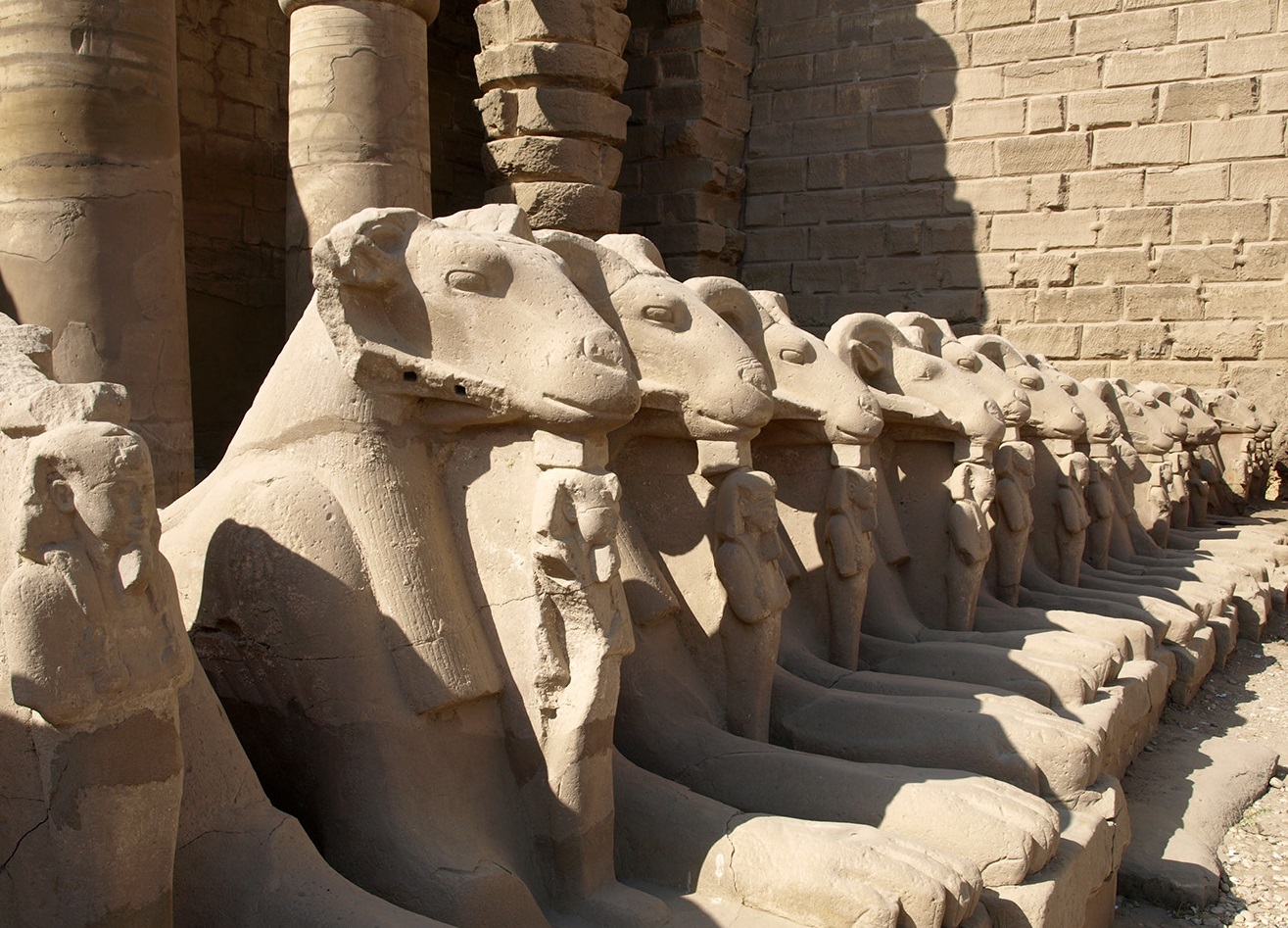 上图:建于主前2000年的古埃及卡纳克神庙(Temple of Karnak)入口的狮身公山羊像,公山羊象征古埃及最高神阿蒙-拉(Amun-Ra),站在公山羊脚前的是法老,象征最高神保护法老。多毛的公山羊是古实(Kush)的主神,埃及征服古实之后,认为古实主神就是太阳神阿蒙-拉,从此公山羊成为阿蒙-拉的化身之一。由于公山羊是多产的象征,所以公山羊形状的阿蒙神又与男性生殖神敏(Min)组合成阿蒙-敏(Amun-Min),进而演化成希腊淫乱的山羊神潘(Pan)。库努牡神(Khnum)也长着一个公羊头。